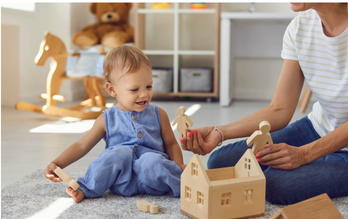 Early childhood educator playing with a toddler using wooden figures and a toy house in an early learning environment.