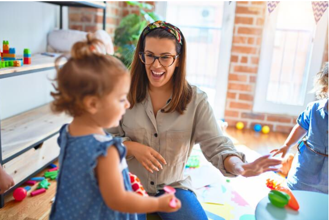 Early childhood educator smiling and interacting with toddlers during a colourful classroom play activity.