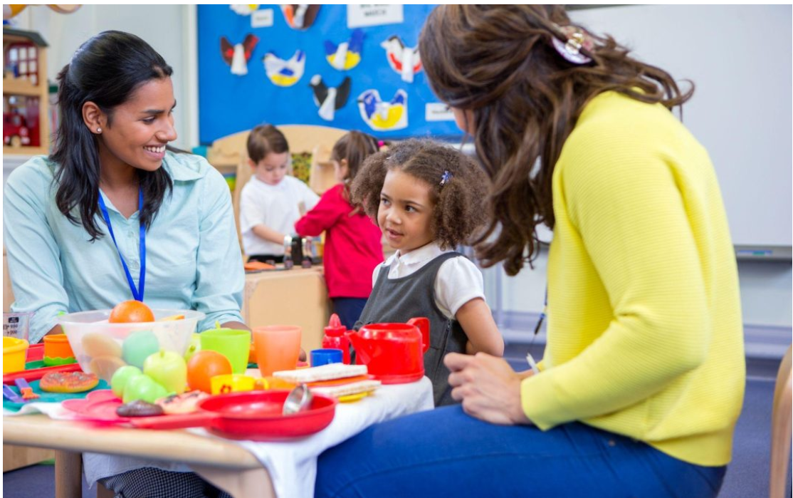 Early childhood educator engaging with a young child during a pretend-play activity in a busy childcare classroom.
