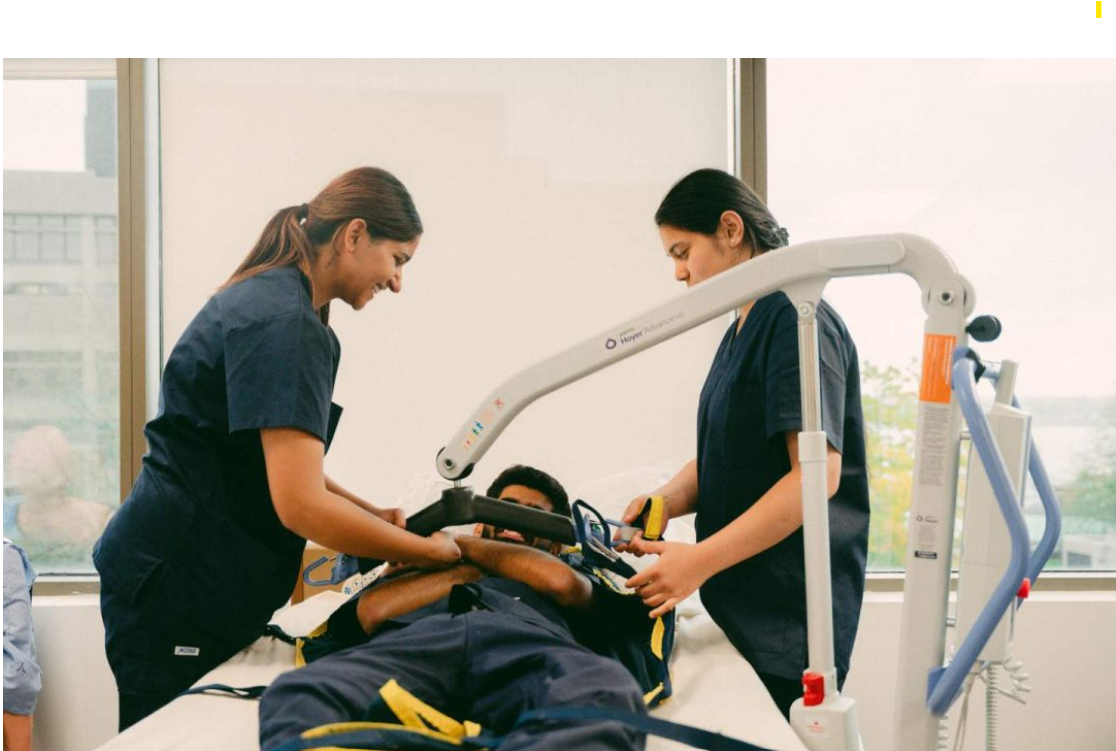 Continuing Care Assistant students practising a safe patient transfer using a mechanical lift in a healthcare training room.