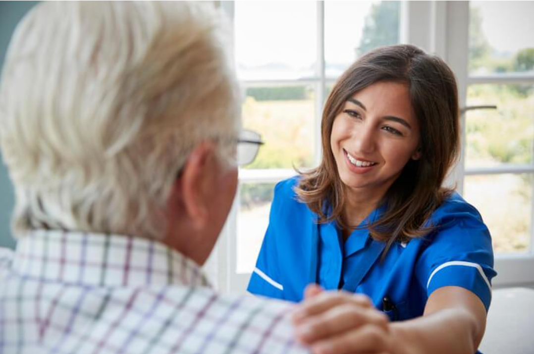 Continuing Care Assistant speaking warmly with an older adult and offering reassurance in a home-care setting.