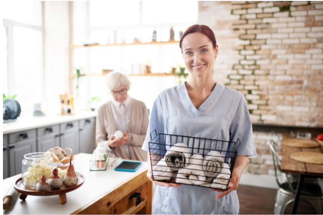 Continuing Care Assistant carrying clean towels while supporting an older adult with daily routines at home.