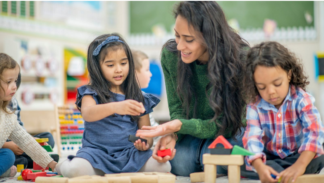 Early childhood educator helping a diverse group of children build structures with wooden blocks in a classroom.