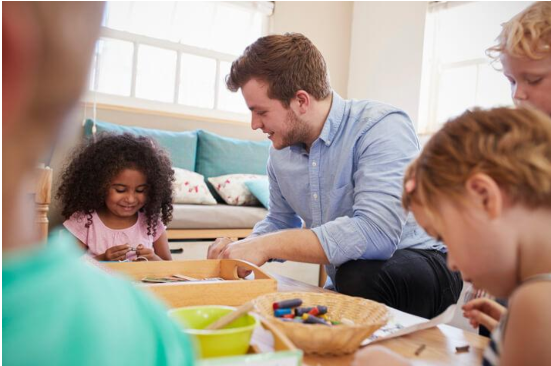Early childhood educator patiently supporting young children during a hands-on craft activity in a childcare classroom.