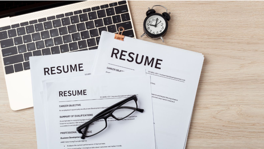 Printed resumes beside a laptop, glasses, and an alarm clock on a desk.