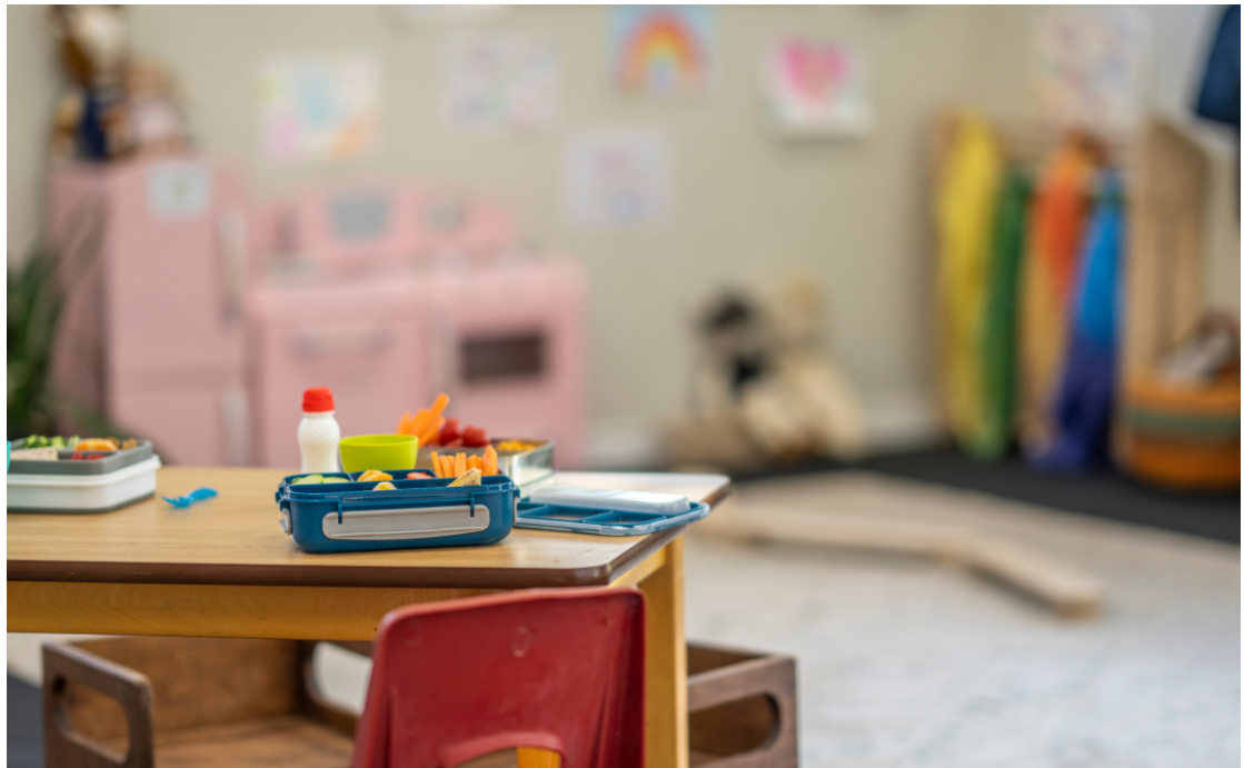 Open lunchbox with healthy snacks on a child-sized table in a bright early learning classroom.